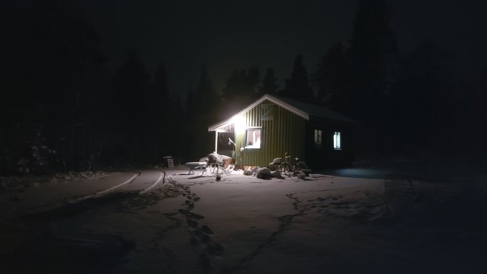 Ett grönt hus upplyst av en lampa i snöigt landskap på kvällen med fotspår i snön.