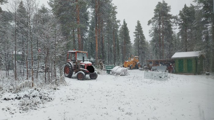 Snöfall över en bondgård med traktorer, skog i bakgrunden och ett grönt skjul.