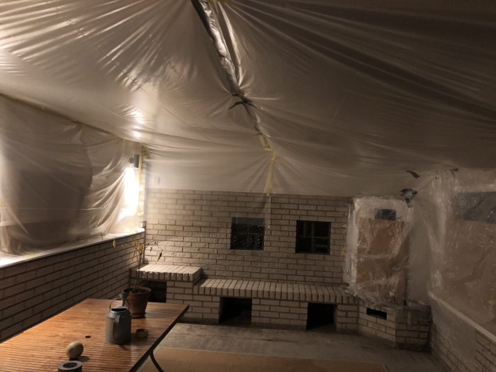Ett under renovering uterum med tegelväggar och plasttäckt tak och möbler, indikerar förberedelse för golvvärmeinstallation.