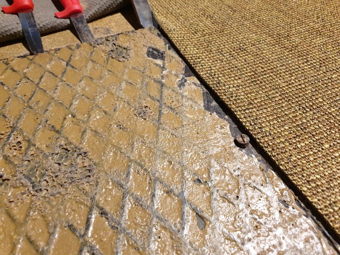 Avlyft hörn av en gulaktig, mönstrad matta avslöjar betongplatta med gammalt lim, möjlig asbestundersökning.