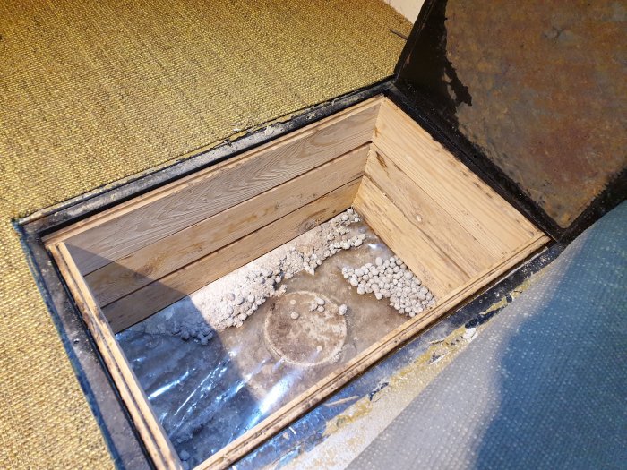 Öppen inspektionslucka i golv med synliga avloppsrör och isoleringsmaterial, bredvid gammal gul matta.