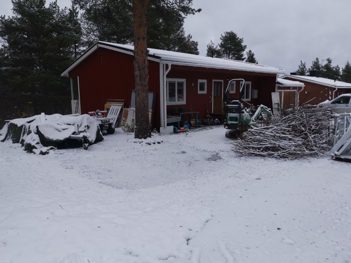 Röd stuga med vita knutar täckt av snö, med en lövhög, uteleksaker och byggmaterial i trädgården.