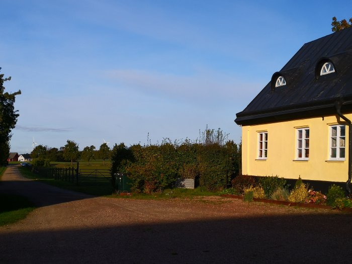 Solig utsikt över ett gult hus med mörkt tak, grusgång och grönskande omgivning som indikerar varmt väder.