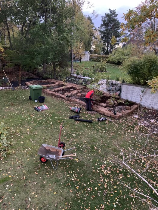 På bilden syns en trädgård med ett pågående markarbetesprojekt som involverar torvblock och löstorv. En person arbetar i det dämrande ljuset.