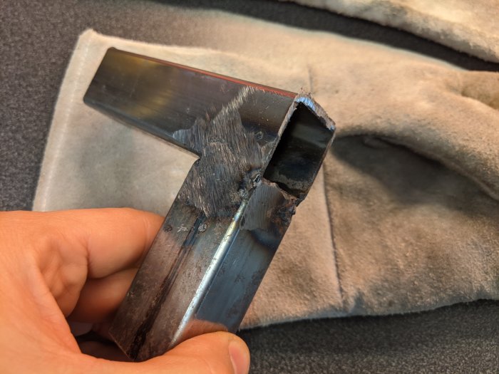 Hand håller en nyligen svetsad metallvinkel med synliga svetsfogar och slipmärken.