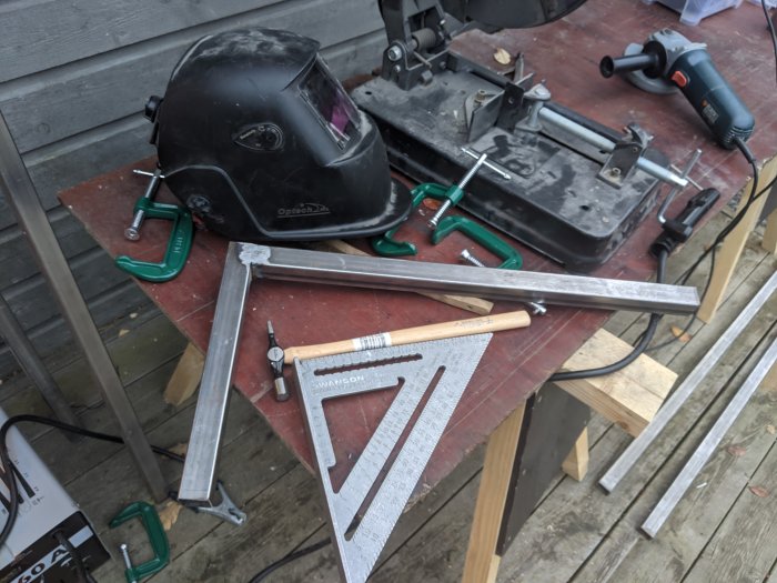 Svetsutrustning och verktyg på arbetsbord inklusive hjälm, vinkelslip och metallvinklar.