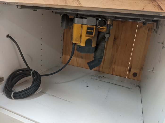 En överhandsfräs av märket DeWalt DW621 monterad under ett fräsbord med synlig elkabel och spånsugskoppling.