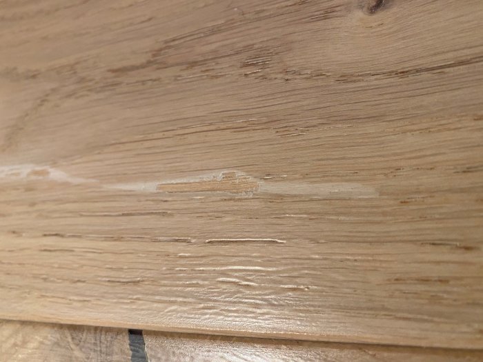 Närbild på en repa på vitpigmenterad mattlackad ekparkettgolv, repans djup på ca 1 mm.