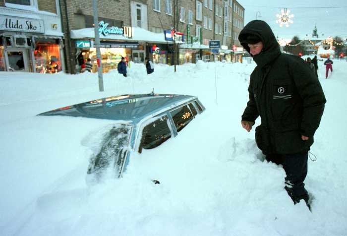 Person tittar på en bil som är nästan helt övertäckt av snö på en stadsgata.