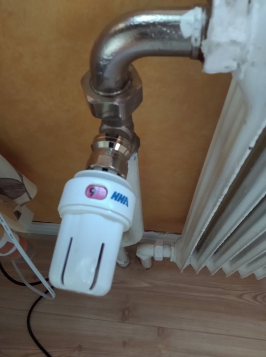 Termostat och ventil på en varm radiator med synliga rör och en kabel på golvet.