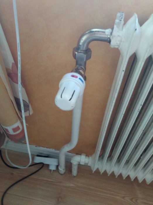 Vit radiator med monterad termostat i ett hushåll, kopplat till VVS-rör, mot en vägg med tapet.