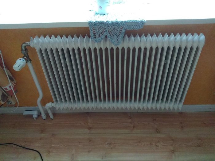 Vit ribbad radiator med termostatvred i ett rum med trägolv och orange vägg, beskriven i VVS-relaterad fråga.