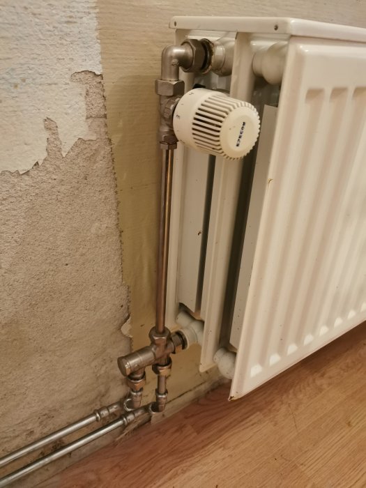 Radiator med termostat och anslutningsrör vid en avskalad vägg.