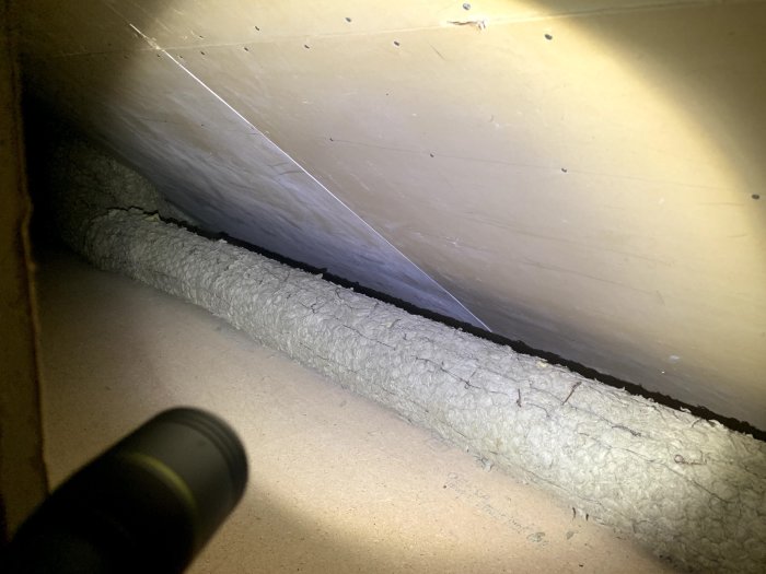 Vy av en vind med isolering längs taket och en mörk glipa där taket möter golvet, belyst med ficklampa.