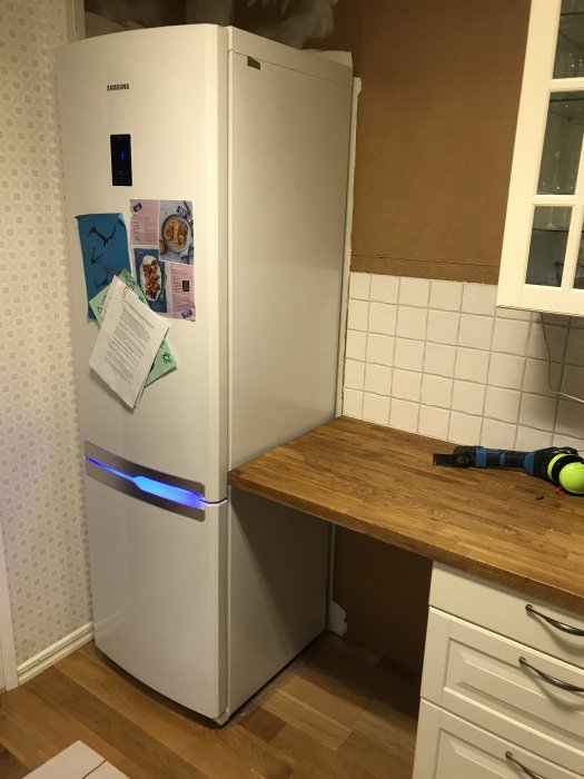 Kök under renovering med vitkaklade väggar, borttagna skåpstommar och synlig kyl och frys.