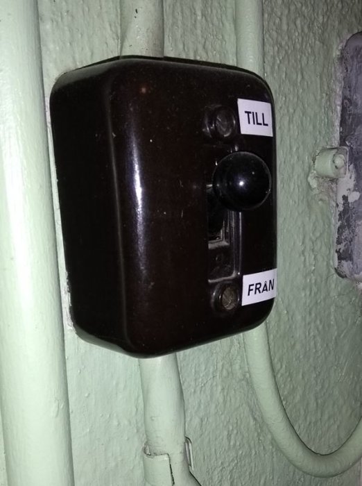 Gammal strömbrytare med etiketter "TILL" och "FRÅN" på en texturerad vägg med rör.