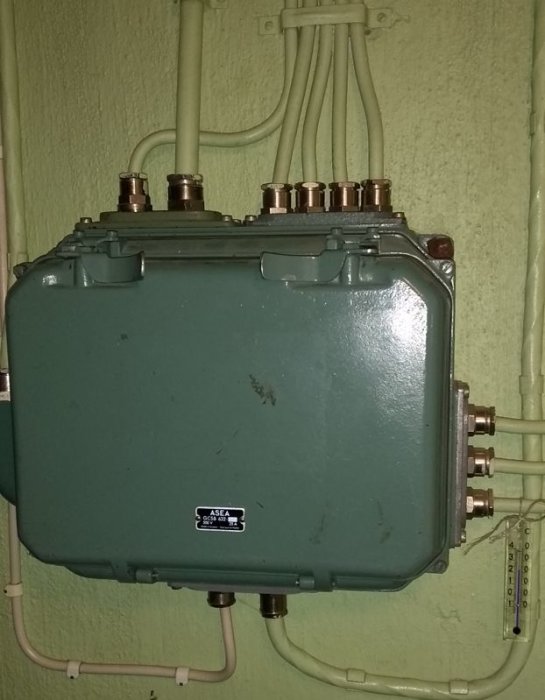 En grön elektrisk fördelningslåda monterad på en vägg med rör och kablar anslutna, märkt med ASEA-logotyp.