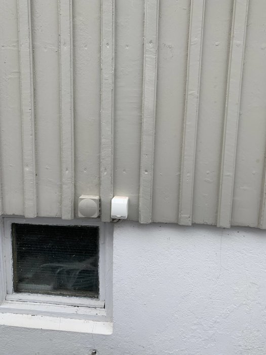 Slutresultatet av en vägg med nyinstallerad vit rörelsedetektor vid en vitplåtad fasad ovanför ett litet fönster.