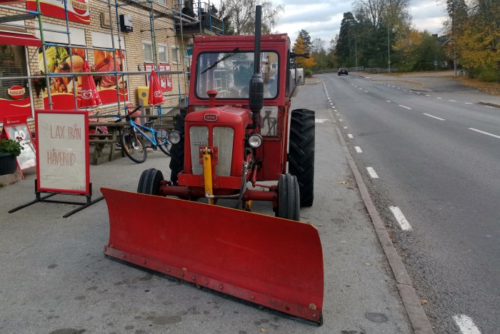 Röd äldre modell traktor med snöblad parkerad vid en vägkant i höstigt landskap.