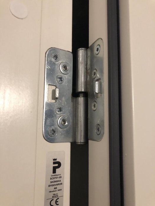 Närbild på ytterdörrsgångjärn med skylt för certifikat på dörrkarmen.