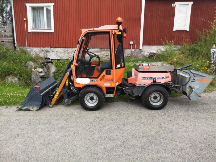 Orange kompaktlastare utrustad med en diagonal snöplog framför en röd stuga.