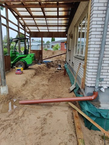 Husdränering under arbete med grävmaskin, dräneringsrör och sand runt husgrund.