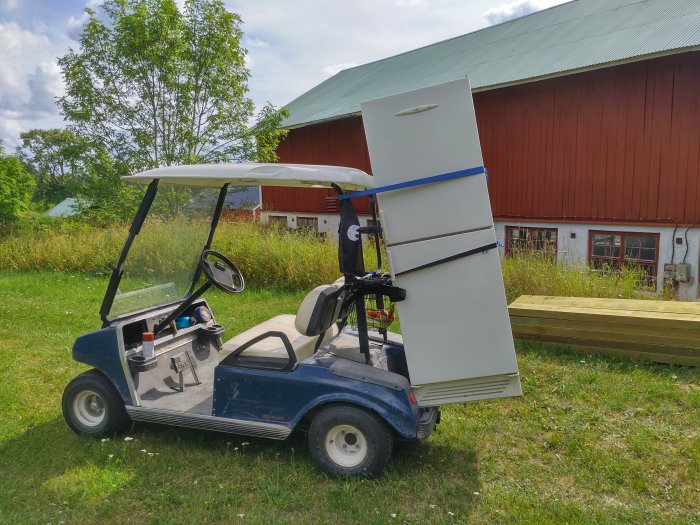 Golfbil med en stor ölkyl fastspänd på sidan framför en röd lada och grönt landskap.