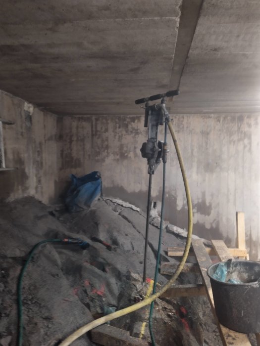 Borrmaskin monterad på borrstång under tak med begränsat utrymme, sladdar och byggmaterial syns.