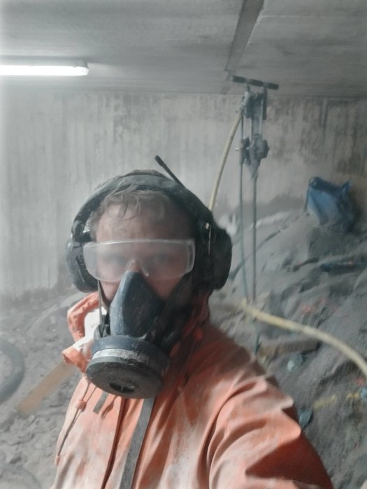 Person i skyddsutrustning tar en selfie under borrningsarbete i en trång arbetsplats.