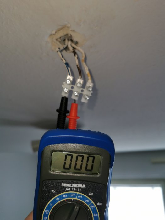 Multimeter visar 0 volt mot en öppen eldosa med flera elkablar i ett tak, indikerar ingen spänning.