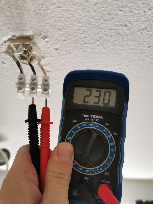Multimeter visar 230 V framför öppen eldosa med blå och gul kabel.
