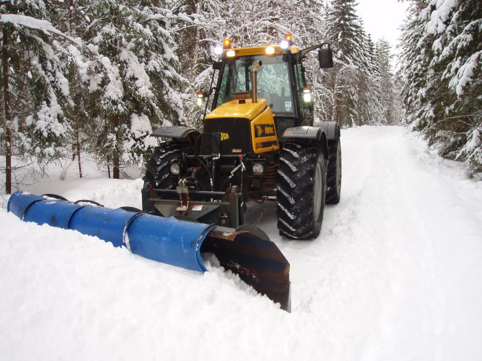 Gul traktor med snöslunga rensar en snötäckt väg omgiven av snötyngda träd.