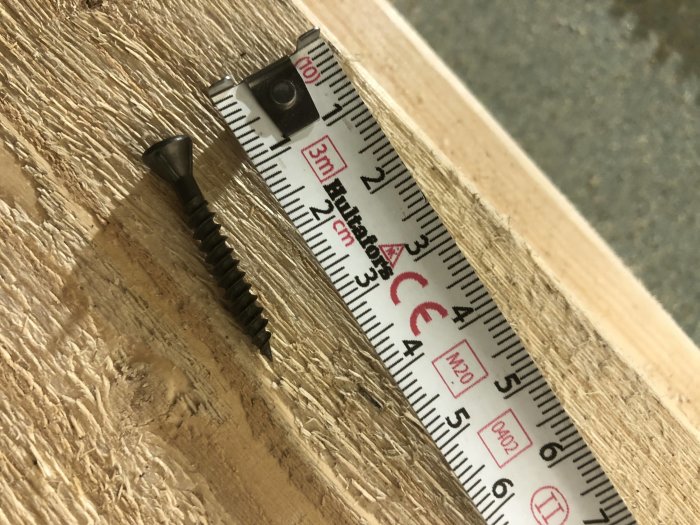 Skruv og målebånd ligger på et stykke træ. Målebåndet viser ca. 5,5 cm. Skruven er sort med forsænket hoved til byggeri.