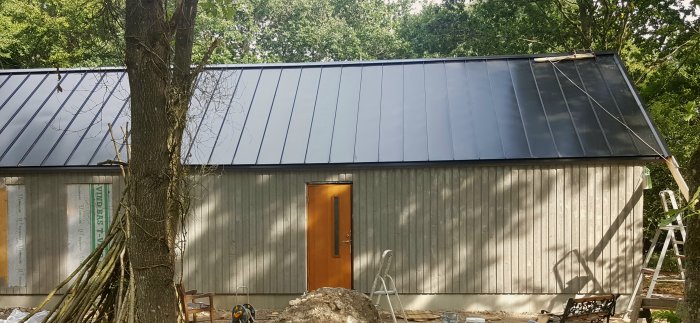 En husfasad under konstruktion med nyinstallerade solpaneler på taket, oavslutad sidobeklädnad och byggmaterial runtomkring.