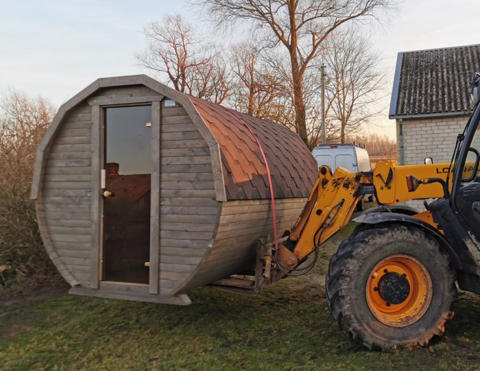 Trähus i tunnelform lyfts av en gaffeltruck, intill ett hus och träd.