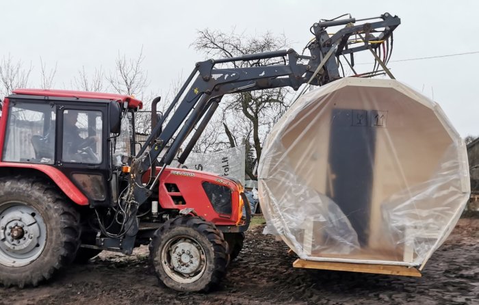 Traktor med frontlastare lyfter en förpackad stor produkt målad i neutral färg.