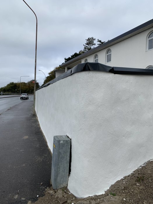 Ojämnt böjd plåtkant längs en vitputsad mur vid en vägkant.