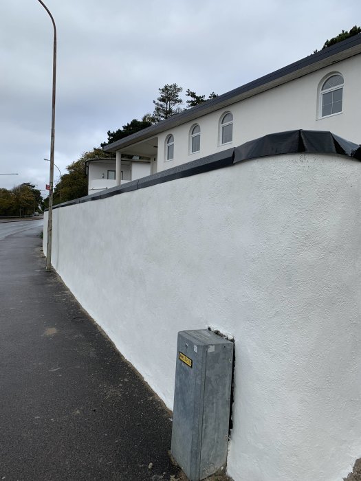 Ojämn böjd plåt på kanten av en vitmurad vägg vid trottoarkanten, mot en grå himmel.
