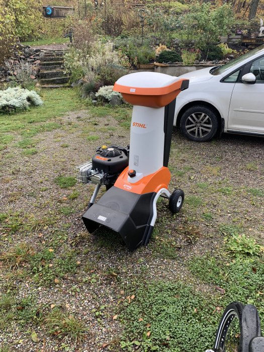 En STIHL trädgårdskvarn parkerad på en grusdrivväg med en lummig trädgård och en vit bil i bakgrunden.