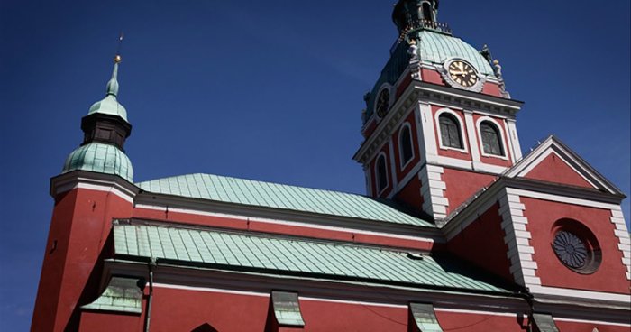 Röd byggnad med grön plåttak, rundade delar på tinnar och ett torn med klocka under blå himmel.