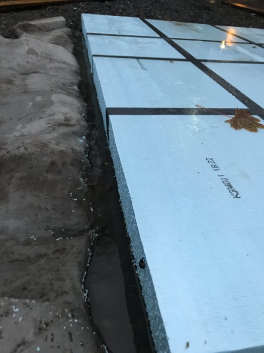 Grävd mark med vattensamling intill rad av XPS-isolering på en uppfart under kvällsbelysning.
