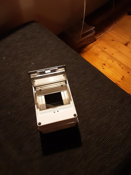 Vit vägguttagslåda ligger på en mörk textilmatta, med en osynlig kabel som sträcker sig ut från bilden.