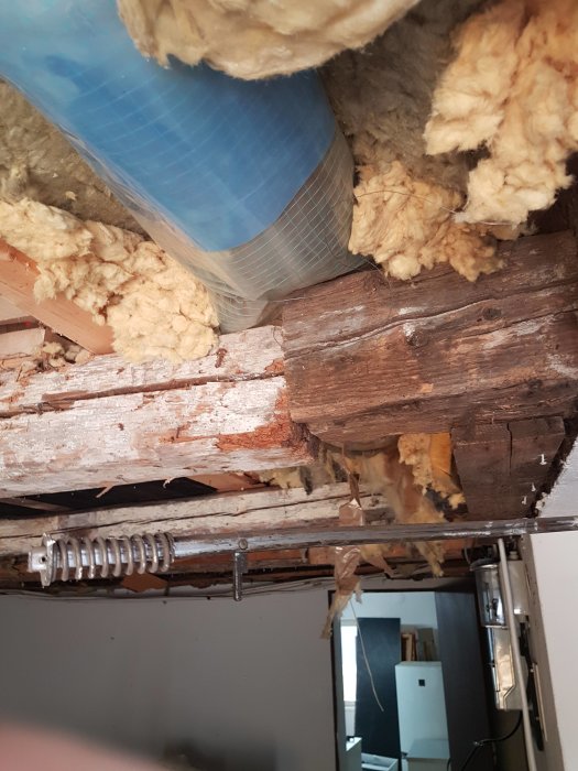 Äldre träbalkar och tillfälliga förstärkningar i ett garage tak med synlig isolering och en genomskärning till rummet nedanför.