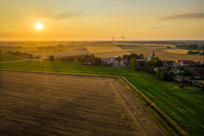 Soluppgång över ett lantligt landskap med fält, vindkraftverk och byggnader i augusti.