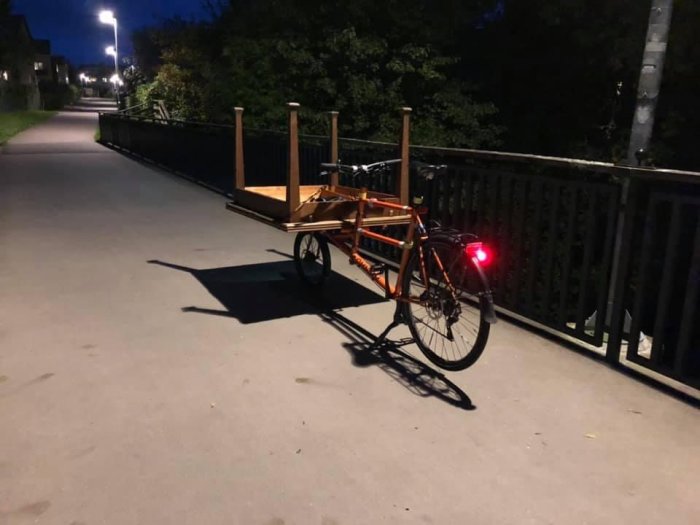 Lång lastcykel med träflak parkerad på en bro vid skymning, lyktor tända.