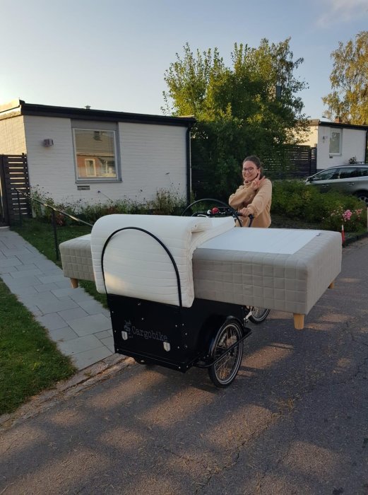 Person transporterar sängmadrass på en lastcykel längs en förortsgata.