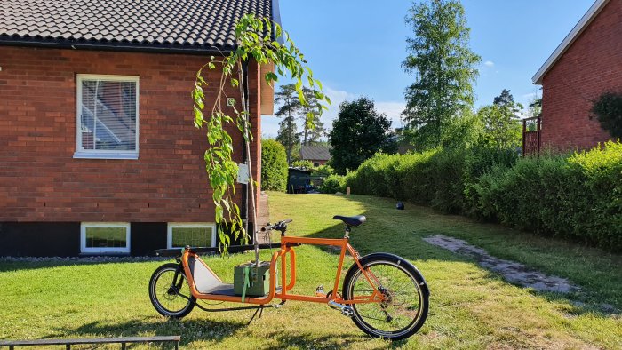 Lastcykel parkerad på gräs framför rött tegelhus, med verktygslåda i lastdelen, på en solig dag.