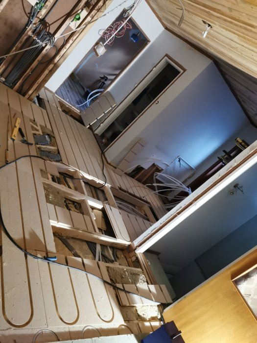 En rörig byggarbetsplats med uppbrutet golv och golvspån som ligger i oordning, kablar och en dörr i bakgrunden.