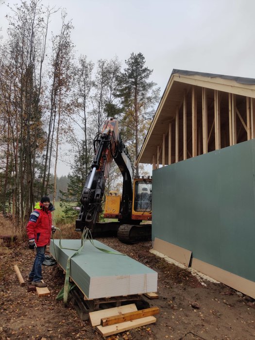 En person står intill en hög med utegips och en grävmaskin vid en husstomme under uppbyggnad.