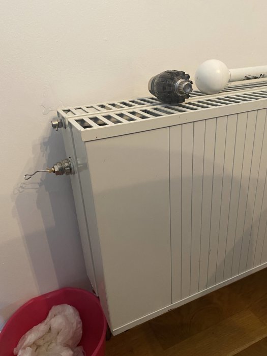 Radiator med mma termostat och okänd insatsventil mot en vit vägg, med en rosa hink nedanför.